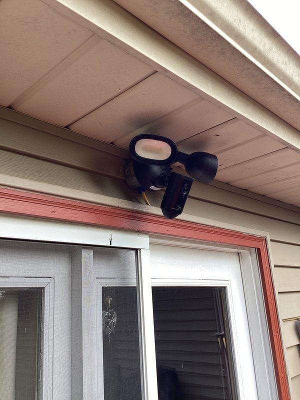 Installed Ring floodlight camera with dual-head motion sensor security lighting on exterior soffit above sliding glass door. Completed electrical bypass box installation to provide proper power supply to the fixture, ensuring full functionality of both the camera system and motion-activated LED floodlights.