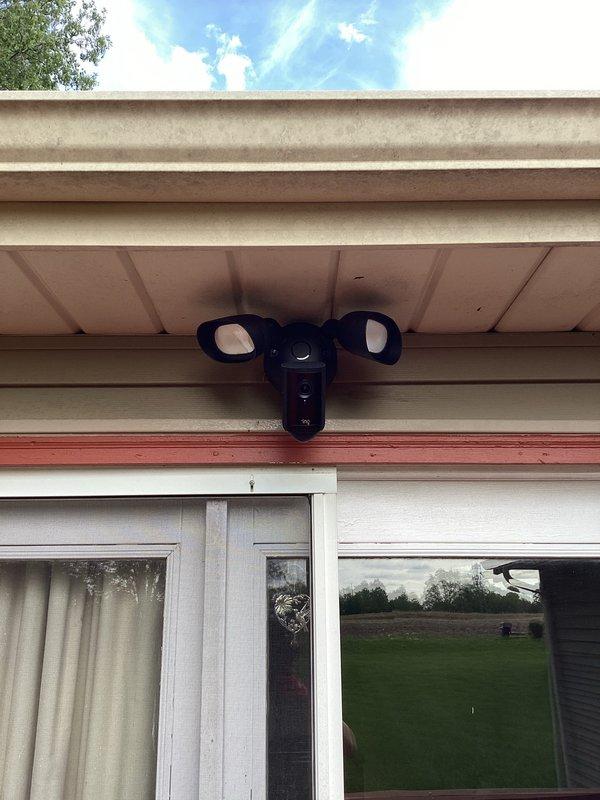 Installed Ring floodlight camera with dual-head motion sensor security lighting on exterior soffit above sliding glass door. Completed electrical bypass box installation to provide proper power supply to the fixture, ensuring full functionality of both the camera system and motion-activated LED floodlights.
