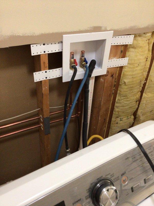 Completed installation of copper supply piping and PVC drainage lines through interior wall cavity for washing machine connection. Installed wall plate with pass-through opening to accommodate red and blue water supply hoses and black corrugated drain hose, ensuring proper routing and secure connections to the appliance. All piping was properly secured with metal strapping and positioned to allow adequate clearance within the wall framing alongside existing insulation.