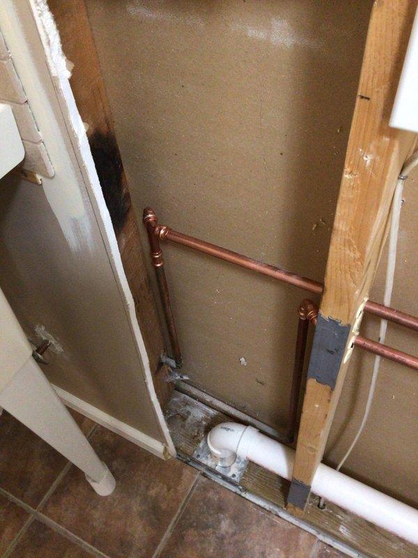 Completed installation of copper supply piping and PVC drainage lines through interior wall cavity for washing machine connection. Installed wall plate with pass-through opening to accommodate red and blue water supply hoses and black corrugated drain hose, ensuring proper routing and secure connections to the appliance. All piping was properly secured with metal strapping and positioned to allow adequate clearance within the wall framing alongside existing insulation.