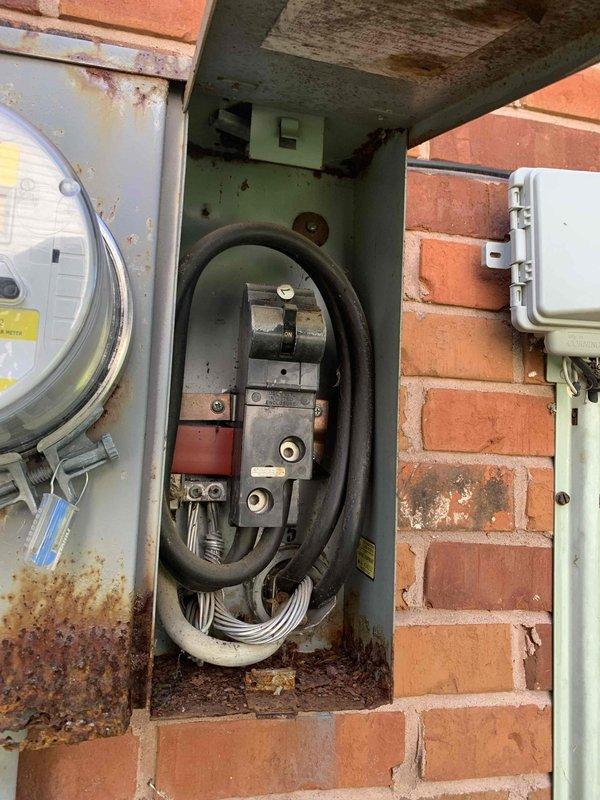 Conducted on-site evaluation of existing outdoor electrical service equipment for meter box replacement estimate. The current installation includes a weathered green metal panel with exposed internal wiring and circuit breakers, an aged utility meter displaying significant rust and corrosion on the housing, and auxiliary weatherproof junction boxes mounted to the brick exterior. Documented the deteriorated condition of the service equipment, noting rust staining around mounting points, accumulated debris in the panel, and water damage indicators on surrounding surfaces. Photographed the complete electrical service configuration including conduit routing, panel positioning relative to other utility equipment, and the overall site layout to provide accurate material and labor estimates for new meter box installation.