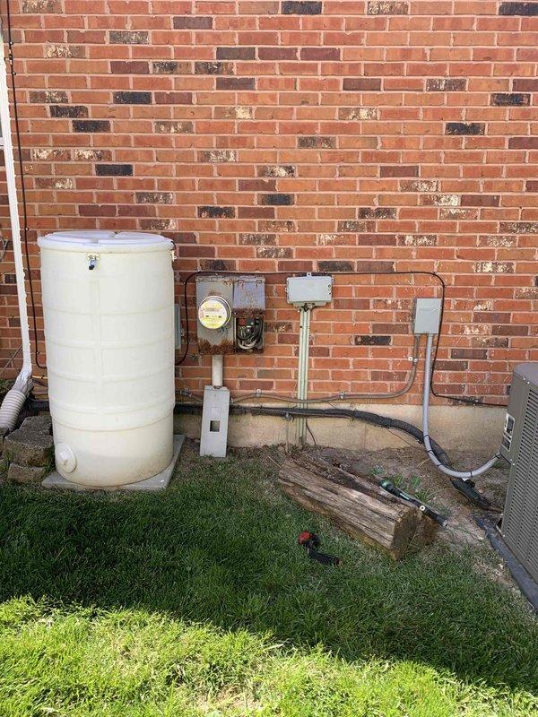 Conducted on-site evaluation of existing outdoor electrical service equipment for meter box replacement estimate. The current installation includes a weathered green metal panel with exposed internal wiring and circuit breakers, an aged utility meter displaying significant rust and corrosion on the housing, and auxiliary weatherproof junction boxes mounted to the brick exterior. Documented the deteriorated condition of the service equipment, noting rust staining around mounting points, accumulated debris in the panel, and water damage indicators on surrounding surfaces. Photographed the complete electrical service configuration including conduit routing, panel positioning relative to other utility equipment, and the overall site layout to provide accurate material and labor estimates for new meter box installation.