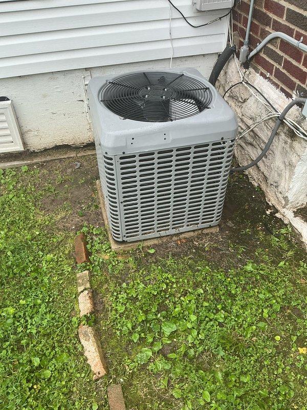 Performed preventative maintenance tune-up on residential outdoor air conditioning condenser unit. Completed standard inspection procedures including checking refrigerant lines, electrical connections, and overall system operation. Unit located on concrete pad against building exterior with vinyl siding, showing normal weathering for outdoor installation. Documented equipment condition with photos showing condenser housing, fan grille assembly, and refrigerant line connections to building. System operational upon completion of maintenance service.