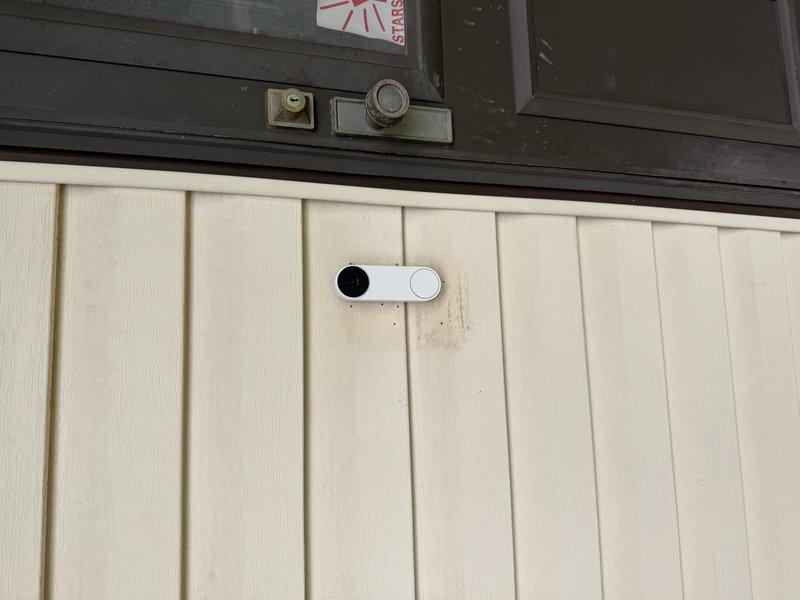 Completed installation of exterior floodlight and video doorbell system. Mounted doorbell camera unit on cream-colored vertical panel siding below dark exterior wall section, ensuring proper positioning for optimal viewing angle and accessibility. Installed floodlight fixture at designated location to provide enhanced exterior illumination and security coverage. Ran necessary electrical wiring, made all required connections per code, and verified proper operation of both fixtures upon completion.