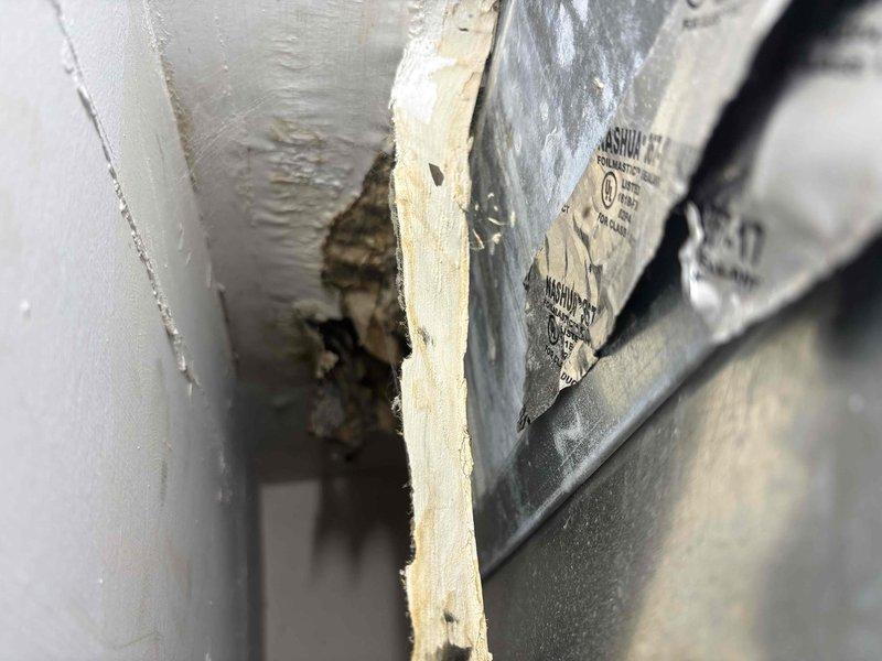 Responded to report of improperly sealed main supply duct on second floor unit installed three months prior. Upon inspection, discovered the main trunk line exiting the air handler was not properly closed off, with opening in drywall/ceiling cavity allowing debris and insulation material to escape into living space. Documented existing conditions showing damaged drywall section with dark debris and insulation visible in cavity, along with exposed ductwork and structural components. The gray supply duct and manufacturer label were visible through the access point, confirming the location of the improperly terminated duct run. Inspection revealed the duct termination required proper sealing and closure to prevent continued debris discharge and maintain system efficiency.