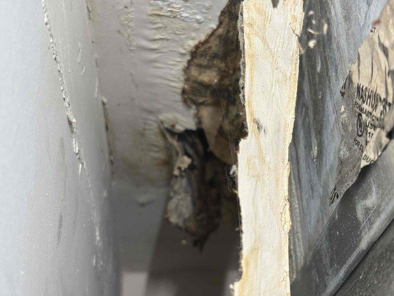 Responded to report of improperly sealed main supply duct on second floor unit installed three months prior. Upon inspection, discovered the main trunk line exiting the air handler was not properly closed off, with opening in drywall/ceiling cavity allowing debris and insulation material to escape into living space. Documented existing conditions showing damaged drywall section with dark debris and insulation visible in cavity, along with exposed ductwork and structural components. The gray supply duct and manufacturer label were visible through the access point, confirming the location of the improperly terminated duct run. Inspection revealed the duct termination required proper sealing and closure to prevent continued debris discharge and maintain system efficiency.
