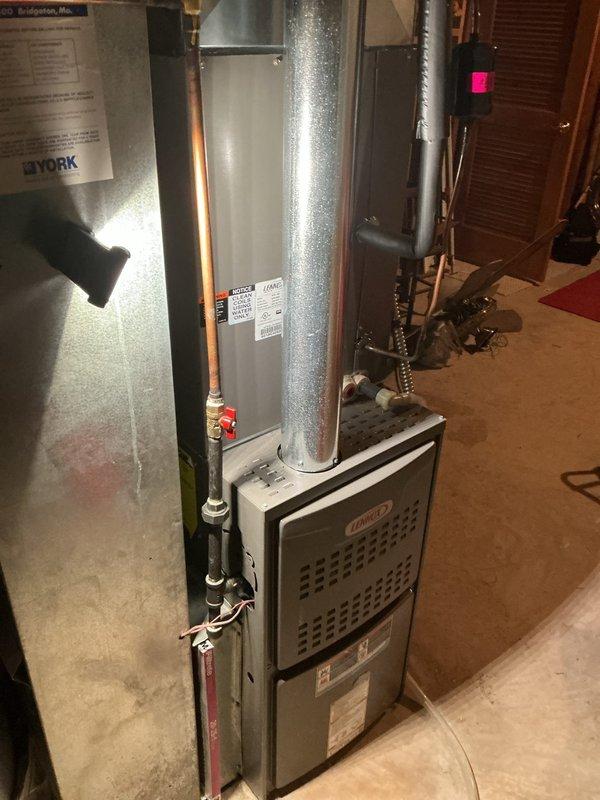 Responded to no cooling complaint on residential HVAC system approximately 10-15 years old. Upon arrival, found entire system inoperative with no power to outdoor condensing unit. Performed diagnostic evaluation of Lennox furnace and air handler assembly located in basement utility area. Inspected electrical connections, refrigerant lines with brass fittings and red-handled service valves, and ductwork configuration. Verified proper installation of copper refrigerant piping and assessed overall system condition. Documented equipment nameplate information and system specifications. Photographed installation including furnace unit, vertical supply ductwork, refrigerant line set, and associated components for service records. System restored to operational status with cooling function confirmed.