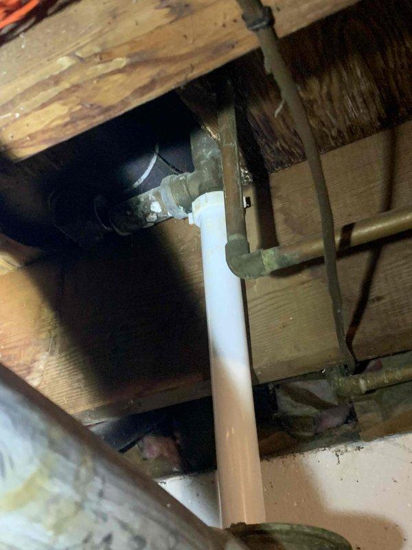 Responded to report of bathtub drain issue with suspected missing drain section beneath upstairs bathroom fixture. Accessed drain assembly from basement ceiling space below bathroom. Upon inspection, identified corroded metal drain elbow fitting connected to white PVC drainage pipe at floor joist level. Metal fitting displayed significant age-related deterioration with greenish patina, indicating potential for leaks or structural compromise. Documented existing conditions of drain assembly, including connection points between metal and PVC components, and surrounding wooden structural elements showing moisture exposure and weathering. Photographed drain configuration through floor opening to record current state of plumbing infrastructure for assessment and repair planning.