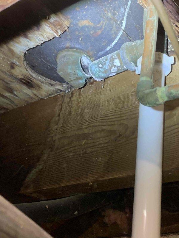 Responded to report of bathtub drain issue with suspected missing drain section beneath upstairs bathroom fixture. Accessed drain assembly from basement ceiling space below bathroom. Upon inspection, identified corroded metal drain elbow fitting connected to white PVC drainage pipe at floor joist level. Metal fitting displayed significant age-related deterioration with greenish patina, indicating potential for leaks or structural compromise. Documented existing conditions of drain assembly, including connection points between metal and PVC components, and surrounding wooden structural elements showing moisture exposure and weathering. Photographed drain configuration through floor opening to record current state of plumbing infrastructure for assessment and repair planning.