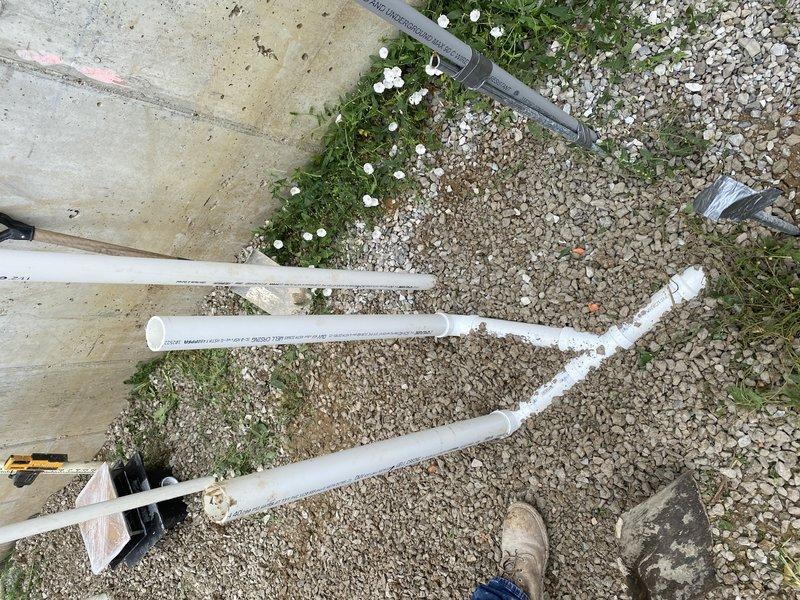 Completed rough-in plumbing installation for outdoor fountain system using white PVC piping configured in a Y-branch layout on gravel surface. Installed drainage piping with elbow fittings connected to collection system near building perimeter, with proper pitch for water flow and condensate management.