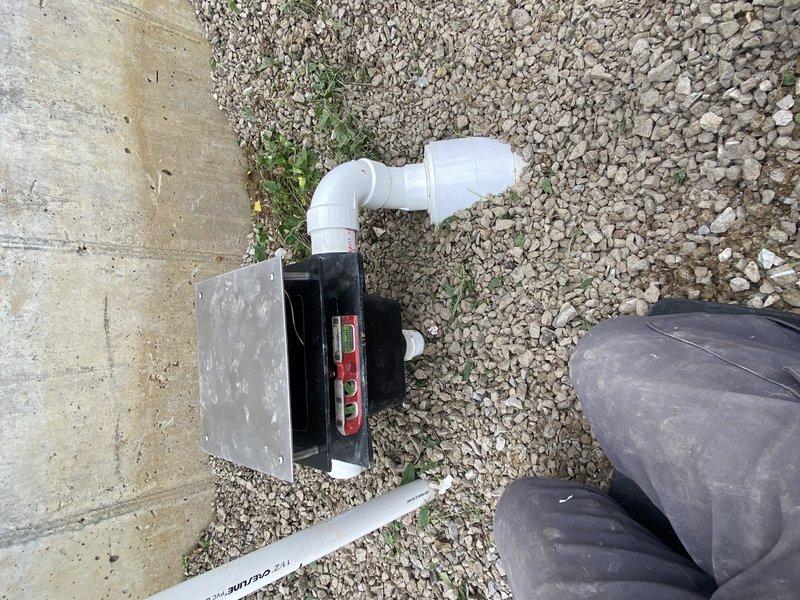 Completed rough-in plumbing installation for outdoor fountain system using white PVC piping configured in a Y-branch layout on gravel surface. Installed drainage piping with elbow fittings connected to collection system near building perimeter, with proper pitch for water flow and condensate management.