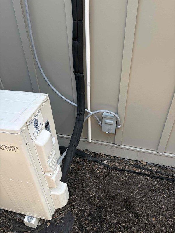 Responded to no cooling complaint on recently installed Mitsubishi Electric mini split system serving living room area. Upon arrival, inspected outdoor condensing unit and refrigerant line set installation mounted on exterior wall. Verified electrical connections at disconnect box and examined conduit routing from outdoor unit. Documented equipment installation with exterior wall-mounted components showing proper line set protection and electrical connections in operational condition.