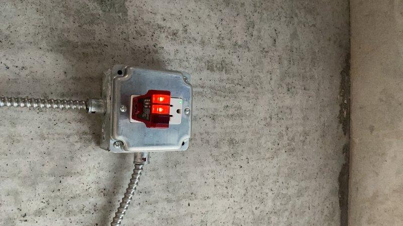 Installed industrial-grade electrical junction box with red receptacle fixture on basement ceiling, utilizing flexible metal conduit for wire protection and routing. Two dedicated conduit runs were secured to the junction box to establish a 20-amp circuit for the newly added outlets, ensuring proper code compliance and safe power distribution to the basement workspace and storage area.