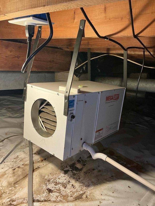 Responded to service call for inspection of two 5-year-old dehumidifiers located in crawl spaces. Accessed both units in confined ground-level areas and performed diagnostic evaluation. Inspected MEGAFLOW dehumidifier unit mounted to floor joists with metal brackets, verified electrical connections, and examined condensate drainage system with PVC piping. Evaluated air intake louvers and assessed overall unit operation. Documented heavily soiled air filter with significant dust and debris accumulation requiring replacement. Checked mounting hardware, ductwork connections, and surrounding crawl space conditions including concrete floor area for moisture concerns.