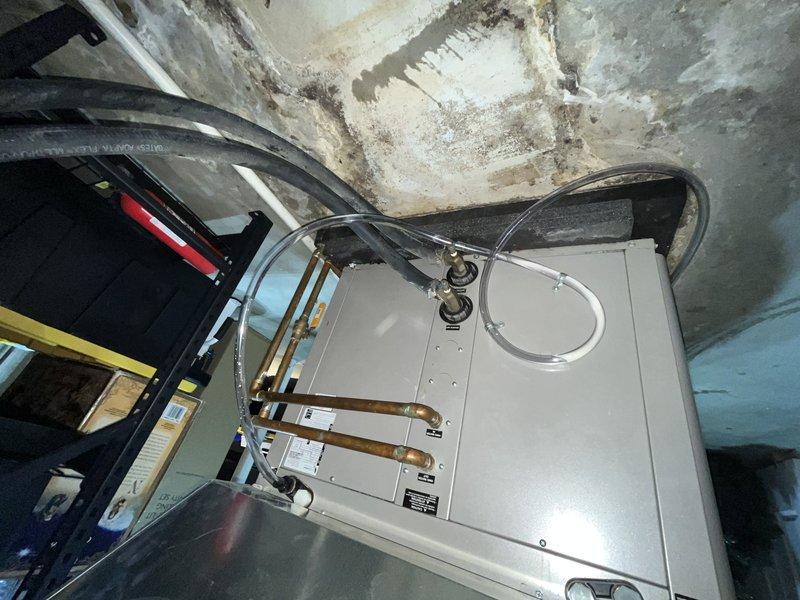 Responded to no cooling complaint on 14-year-old GeoComfort geothermal heat pump system. Upon arrival, confirmed air handler was operating and circulating air through ductwork, but system was not producing cool air. Performed diagnostic evaluation of ground source heat pump components. Inspected air handler unit located in mechanical room, checked refrigerant lines, and verified electrical connections. Documented equipment condition and installation environment showing age-related wear in utility space. System evaluation revealed cooling malfunction requiring further assessment of compressor operation and refrigerant circuit. Photographed equipment for service records showing air handler cabinet and copper refrigerant line connections in basement installation area with visible moisture exposure patterns on surrounding surfaces.