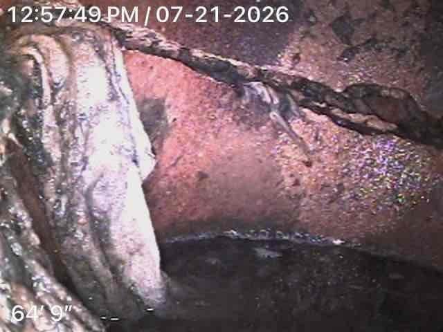 Performed video camera inspection of main sewer line to investigate reported backup issues at rear drain. Camera inspection revealed significant deterioration and corrosion of pipe interior at approximately 64 feet 9 inches from access point, with visible mineral deposits, scaling, and compromised pipe integrity. Documented conditions including standing water, corroded metal surfaces, and structural degradation throughout the affected section. Captured photographic evidence of exterior access point and internal pipe conditions for further evaluation and follow-up repair recommendations.