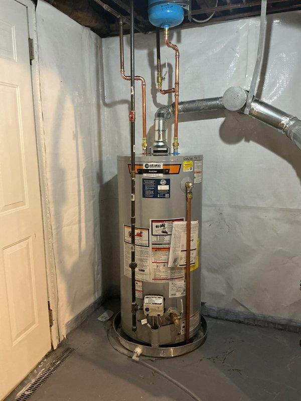 Completed installation of 40-gallon gas tall water heater under permit #26PLU-14536. Installed Ruud brand tank-style unit on metal drip pan with proper clearances in basement utility area. Connected copper supply lines, installed blue expansion tank on ceiling joists, and routed temperature/pressure relief valve discharge pipe per code requirements. Configured gas piping connections and verified all plumbing connections for proper operation. Applied required plumbing identification sticker to completed installation.