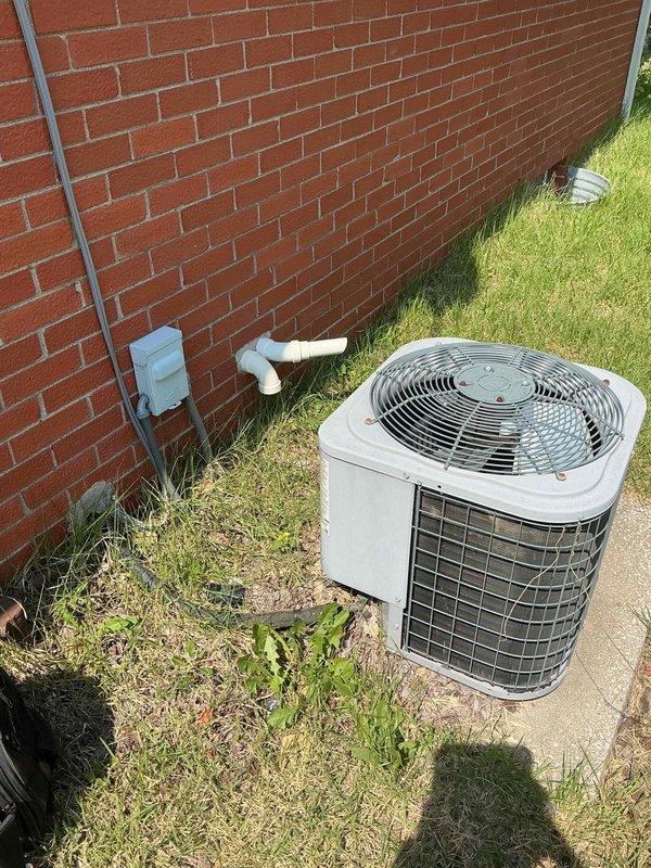 Responded to no cooling complaint on 9-year-old residential AC unit where condenser fan was running but system failed to produce cold air, with intermittent 2-second humming occurring every few minutes. Upon arrival, performed diagnostic evaluation of outdoor condenser unit. Removed access panel to inspect electrical components and wiring. Conducted electrical testing using multimeter to assess capacitor functionality and contactor operation. Photographed unit exterior showing condenser positioned on concrete pad against brick wall with condensate drain line, and documented internal electrical compartment revealing capacitor, contactor, and wire connections during diagnostic process.