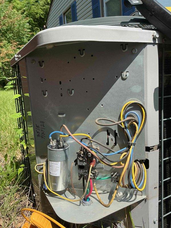 Responded to no cooling complaint on 9-year-old residential AC unit where condenser fan was running but system failed to produce cold air, with intermittent 2-second humming occurring every few minutes. Upon arrival, performed diagnostic evaluation of outdoor condenser unit. Removed access panel to inspect electrical components and wiring. Conducted electrical testing using multimeter to assess capacitor functionality and contactor operation. Photographed unit exterior showing condenser positioned on concrete pad against brick wall with condensate drain line, and documented internal electrical compartment revealing capacitor, contactor, and wire connections during diagnostic process.