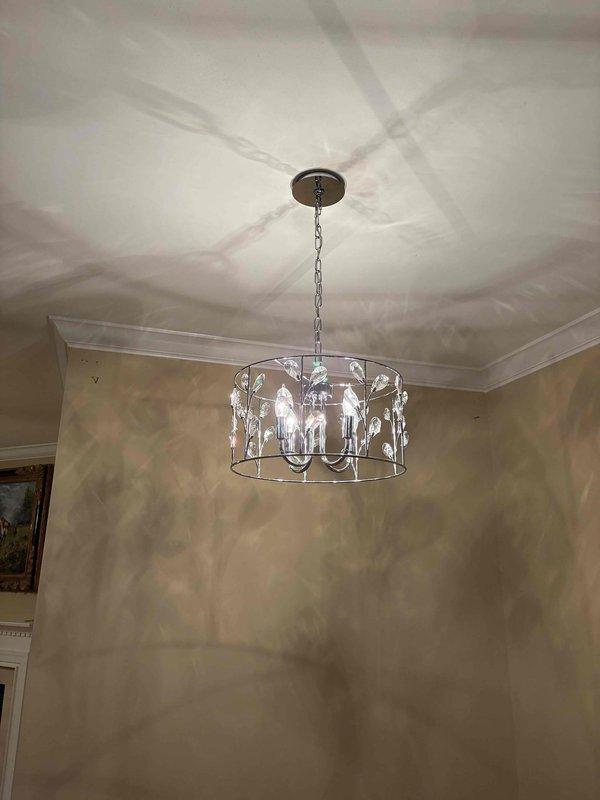 Installed decorative bronze chandelier with crystal accents in residential interior space, featuring cylindrical metal frame with leaf motif details and multiple candelabra-style bulbs. Secured fixture to ceiling mount with chain suspension system, verified proper electrical connections, and confirmed all lighting elements functioning correctly upon completion.