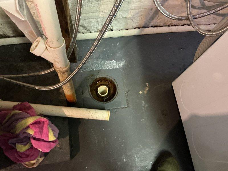Responded to follow-up service call for washing machine drain backing up and overflowing into basement floor drain. Previous technician was unable to clear blockage and suspected cracked floor drain trap. Attempted to clear obstruction using mechanical drain cleaning methods but was unsuccessful in opening the line. Upon inspection, confirmed that the floor drain trap assembly appears to be compromised and requires replacement rather than standard drain cleaning. Documented existing conditions with photographs showing floor drain configuration, surrounding PVC drainage piping, and water heater connections in utility area. Recommended scheduling replacement bid for new floor drain trap installation to resolve ongoing drainage issue.