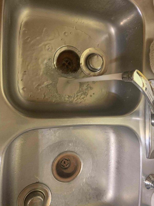 Responded to service call for kitchen sink backing up with clear liquid and draining slowly. Upon arrival, inspected double-basin stainless steel sink with garbage disposal system. Found drainage issue affecting primary basin with disposal unit. Assessed plumbing configuration, including drain assemblies, strainer baskets, and disposal connections. Documented existing conditions showing mineral deposits and residue buildup around drain openings. Cleared obstruction in drainage system to restore proper flow and functionality to kitchen sink.