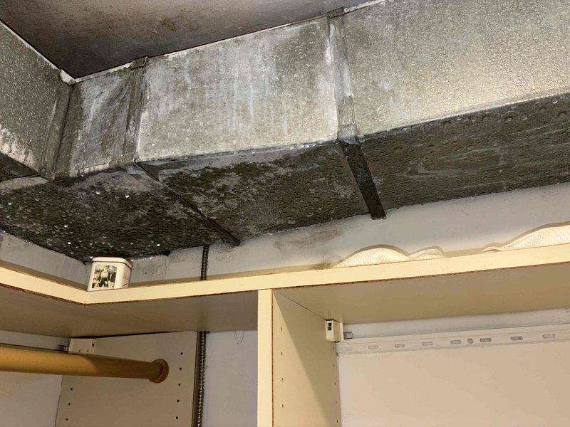 Responded to report of condensation and mildew growth in bedroom closet. Upon inspection, discovered extensive mold and moisture staining on exposed concrete surfaces above closet shelving, with dark discoloration indicating prolonged moisture exposure. Identified large rectangular supply duct running through closet space as probable source of condensation issues. Duct insulation appeared inadequate or compromised, allowing cold surface temperatures to contact warm, humid air and create condensation conditions. Documented extensive mold growth patterns on ceiling and wall surfaces surrounding ductwork. Recommended immediate remediation of mold contamination, proper sealing and insulation of ductwork to eliminate condensation, and verification of proper system operation including airflow balance and humidity control. Advised that both HVAC units should be evaluated for proper dehumidification performance to prevent recurrence.