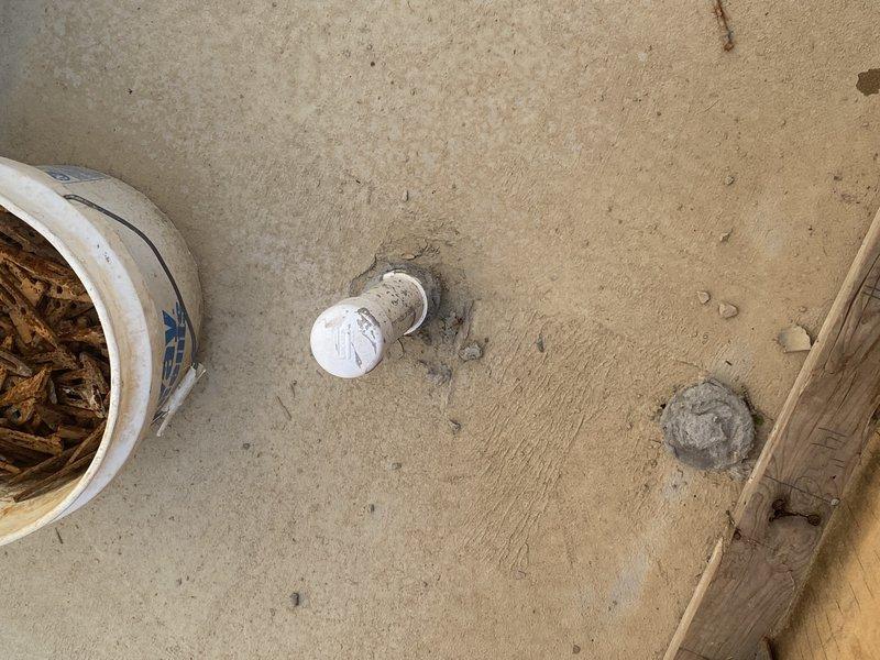 Conducted plumbing site inspection and documented existing conditions. Photographed deteriorated wall-mounted access panel showing significant rust damage, paint failure, and structural compromise with exposed cavity at damaged edge. Documented PVC pipe connection and mounting hardware condition. Also photographed floor-level pipe stub or drain opening protruding from concrete surface, along with visible floor impressions indicating previous fixture or equipment placement. Images captured for assessment of required repairs to damaged access panel and evaluation of rough-in plumbing conditions.