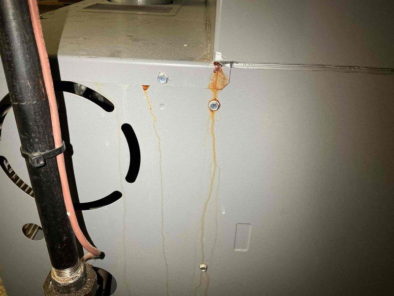 Responded to reported water leak from basement furnace unit over 10 years old. Upon inspection, identified significant rust staining and corrosion on furnace cabinet panel and evaporator coil assembly, indicating chronic moisture exposure. Documented extensive rust deposits on heat exchanger fins, refrigerant line connections, and metal housing components. Photographed affected areas showing corrosion patterns originating from mounting points and spreading across cabinet surfaces, as well as deterioration along coil edges and copper refrigerant lines.
