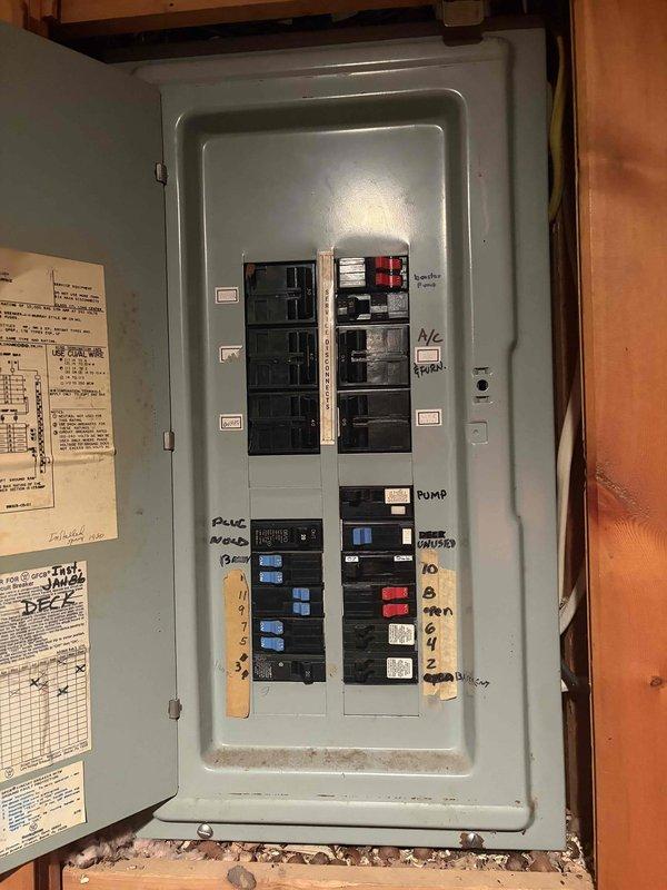 Completed comprehensive Elite electrical inspection of residential service panel and distribution system. Documented existing main breaker panel installation in wooden-framed utility area, noting standard gray metal enclosure with hinged door and secure latch mechanism. Inspected interior circuit breaker configuration revealing multiple branch circuits with mixed breaker types including standard single-pole and multi-pole devices. Observed existing circuit identification system with handwritten labels and documentation posted adjacent to panel for reference. Verified panel accessibility and noted presence of service documentation including circuit schedules affixed to surrounding wall structure. Assessed overall panel condition, identifying normal operational wear and minor surface soiling consistent with typical residential use. Photographed panel exterior and interior arrangements for maintenance records, capturing current breaker positions, labeling system, and general installation 