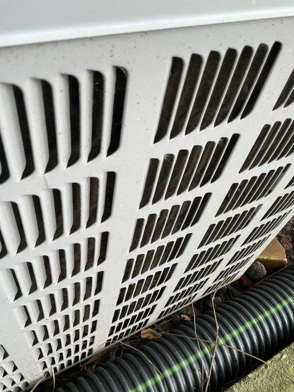 Responded to no cooling complaint on 10+ year old outdoor AC unit. Upon arrival, confirmed unit was blowing warm air with rising indoor temperatures. Inspected exterior condensing unit located at side of home and identified blocked foundation ventilation grates restricting proper airflow to equipment. Removed accumulated debris, leaves, mulch, and organic matter from vent openings and surrounding area. Cleared drainage hose obstruction and restored adequate ventilation clearance around unit foundation. Verified airflow restoration and advised on maintaining clear perimeter for optimal system performance.