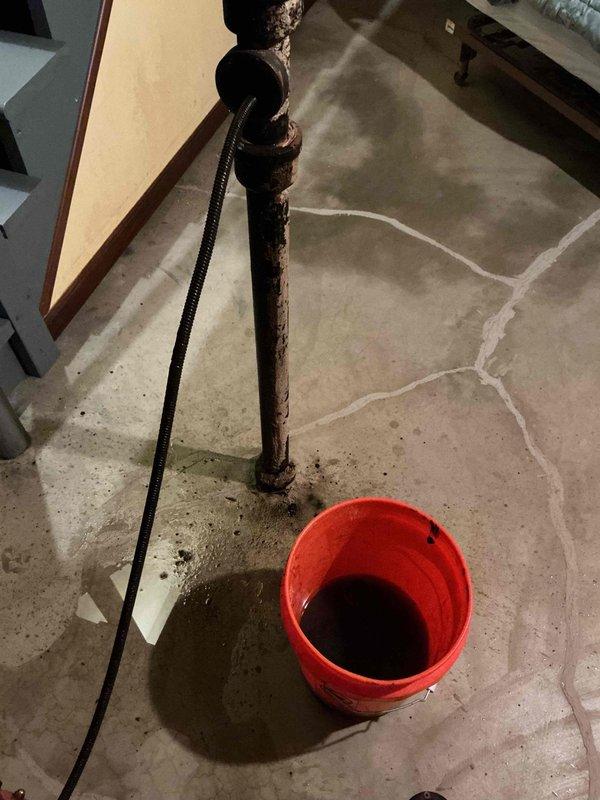 Responded to kitchen sink clog on main living level. Upon arrival, discovered the drainage issue was related to severely corroded vertical plumbing stack visible in lower level beneath kitchen. The deteriorated metal pipe showed extensive rust and corrosion throughout, with active water leakage at base requiring bucket placement for containment. Assessed pipe condition and cleared obstruction in drainage system to restore proper flow. Documented extensive corrosion damage to vertical stack for future repair recommendations, as pipe replacement will be necessary to prevent continued leakage and potential water damage to surrounding areas.