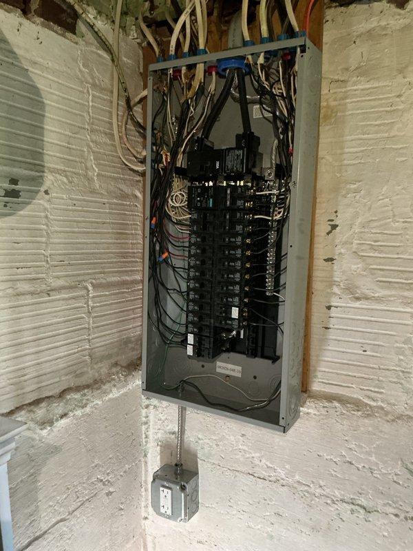 Completed electrical panel installation in residential basement utility area. Installed new gray electrical panel box on white painted brick wall with proper mounting and code-compliant wire management. Configured dual vertical rows of circuit breakers within panel enclosure. Terminated multiple black electrical cables through blue wire clamp connector at panel top, along with tan-colored conduit runs extending along wall and ceiling joists. Installed metal conduit pipe from panel to lower wall-mounted junction box. Routed necessary wiring through exposed ceiling joists to integrate with existing electrical infrastructure. Verified all connections, tested circuit breaker functionality, and ensured installation meets current electrical code requirements for residential service.