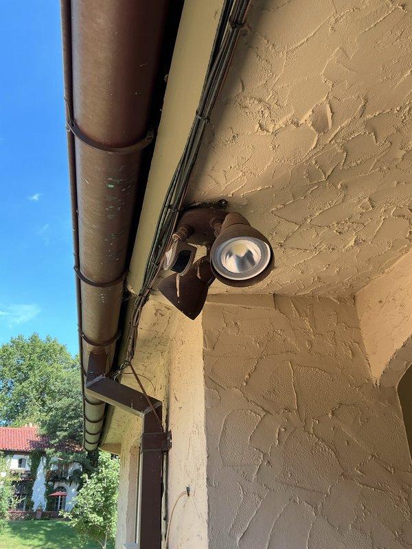 Replaced failed exterior motion detector light fixture at driveway location with customer-supplied dual-head security light. Removed existing 12-year-old single fixture, verified electrical connections, and installed new motion-sensor lighting unit on textured stucco wall beneath eave and gutter system. All wiring connections secured and fixture tested for proper operation.