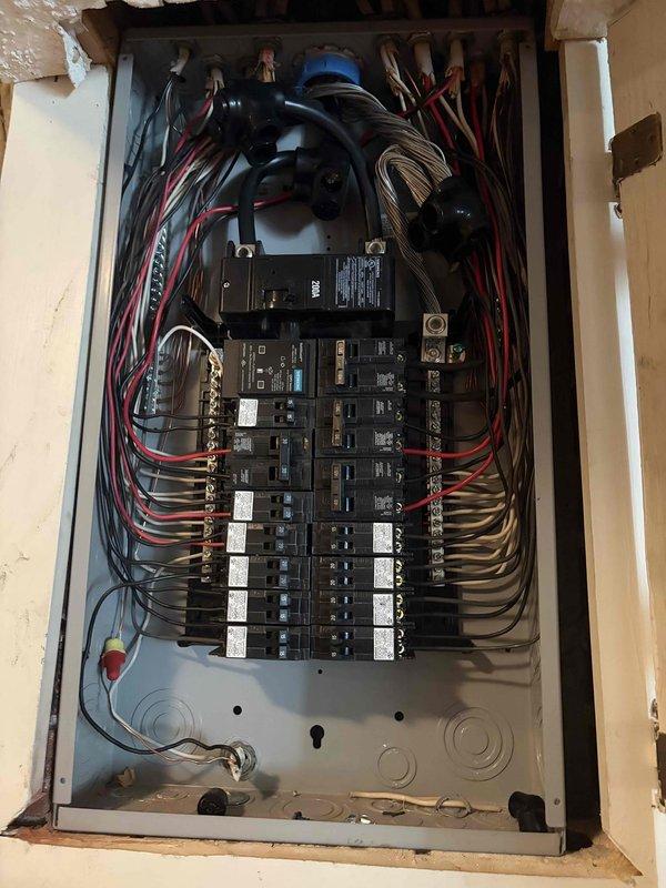 Completed installation of new electrical service panel in residential interior space during construction phase. Panel features dual rows of circuit breakers with proper wire management, including black, red, and tan conductors organized in symmetrical routing from top entry point. Installed main disconnect device, connected all branch circuits to breakers with appropriate labeling, and ensured proper termination at bus bars and terminal connections. All wiring secured according to code requirements with neat curved runs along panel sides.