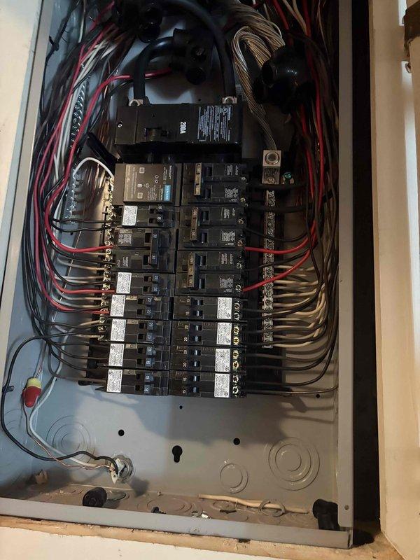 Completed installation of new electrical service panel in residential interior space during construction phase. Panel features dual rows of circuit breakers with proper wire management, including black, red, and tan conductors organized in symmetrical routing from top entry point. Installed main disconnect device, connected all branch circuits to breakers with appropriate labeling, and ensured proper termination at bus bars and terminal connections. All wiring secured according to code requirements with neat curved runs along panel sides.