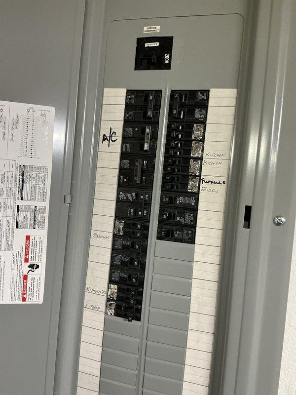 Completed comprehensive electrical inspection of residential breaker panel and associated wiring systems. Documented panel configuration showing dual-column arrangement of circuit breakers with proper labeling for A/C, kitchen circuits, furnace, and garage. Verified main disconnect switch operational status and confirmed all breakers properly seated. Inspected exposed wiring runs above panel, including bundled conductors routing through ceiling structure to blue junction box mounted on structural framing. Panel exhibited clean condition with no visible corrosion, damage, or safety concerns. All manufacturer documentation present on interior door for reference.