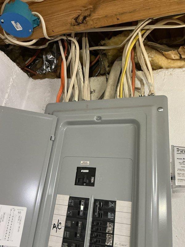 Completed comprehensive electrical inspection of residential breaker panel and associated wiring systems. Documented panel configuration showing dual-column arrangement of circuit breakers with proper labeling for A/C, kitchen circuits, furnace, and garage. Verified main disconnect switch operational status and confirmed all breakers properly seated. Inspected exposed wiring runs above panel, including bundled conductors routing through ceiling structure to blue junction box mounted on structural framing. Panel exhibited clean condition with no visible corrosion, damage, or safety concerns. All manufacturer documentation present on interior door for reference.