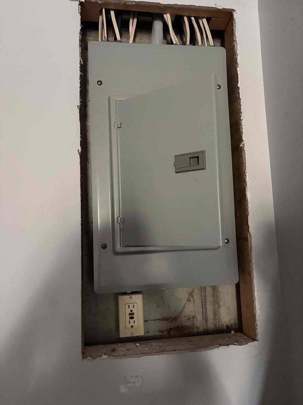Performed comprehensive electrical estimate for replacement of existing 30-year-old main electrical panel and failed breaker. Inspected interior recessed panel installation showing light gray hinged metal cover with multiple feed wires entering from top, noted exposed framing around wall cutout and receptacle below panel. Evaluated exterior electrical disconnect box mounted on building exterior serving HVAC equipment. Documented current panel condition, photographed all components, and provided detailed replacement recommendations for upgraded electrical service panel to meet current code requirements and restore safe operation.