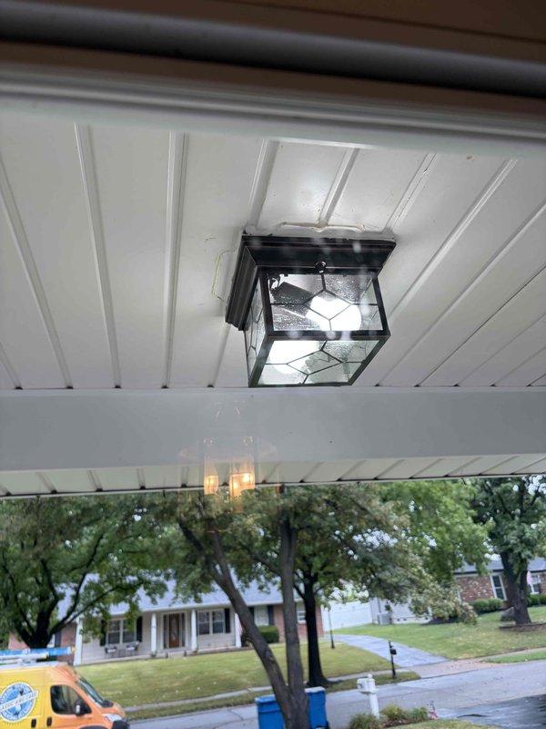Installed customer-supplied decorative outdoor light fixture on covered porch ceiling. The black metal lantern-style fixture with diamond-pattern glass panels was successfully mounted to white beadboard ceiling and tested for proper operation.