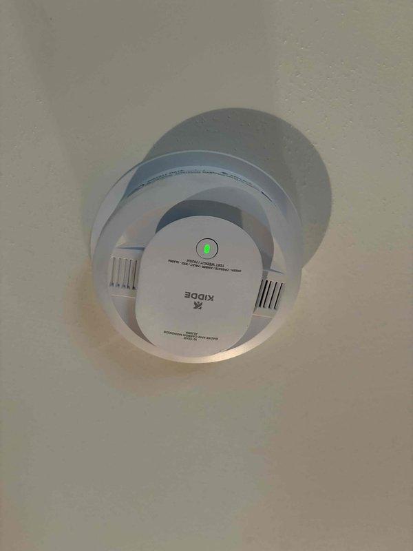 Responded to service call for smoke alarm activation on vaulted ceiling. Inspected malfunctioning Kidde smoke detector using extension ladder to access elevated mounting location. Performed comprehensive inspection of all smoke detectors throughout residence to verify proper operation, mounting integrity, and battery status. Removed and tested the problematic unit, confirming proper sensor function and LED indicator operation. All smoke alarms in home were found to be operational with no additional deficiencies noted.