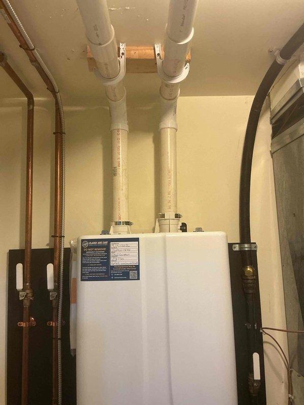 Completed installation of Energy Star certified tankless water heater system in utility room with proper venting, pressure tank integration, and condensate management. Installed all required copper supply piping with corrugated flexible connectors, PVC venting system extending through ceiling with secure mounting brackets and wooden support, and metallic condensate drain pan beneath unit. Secured all piping with appropriate black pipe straps and wall-mounted blue pressure tank for system operation. Applied permit sticker per permit #26PLU-18741 under license #P8730. All connections tested and system commissioned for proper operation.