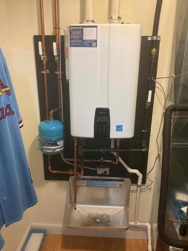 Completed installation of Energy Star certified tankless water heater system in utility room with proper venting, pressure tank integration, and condensate management. Installed all required copper supply piping with corrugated flexible connectors, PVC venting system extending through ceiling with secure mounting brackets and wooden support, and metallic condensate drain pan beneath unit. Secured all piping with appropriate black pipe straps and wall-mounted blue pressure tank for system operation. Applied permit sticker per permit #26PLU-18741 under license #P8730. All connections tested and system commissioned for proper operation.