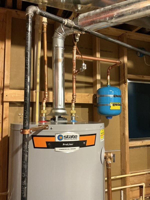 Completed professional installation of a 50-gallon tall gas water heater in an unfinished basement utility area. The State ProLine Commercial Grade unit was installed with comprehensive plumbing connections, including copper supply lines, brass fittings, multiple shut-off valves with color-coded handles for system control, and proper venting with gray flue piping. A blue thermal expansion tank was mounted to the system and secured with copper piping to protect against pressure buildup. All connections were properly fitted and sealed to manufacturer specifications, with the unit positioned on a concrete floor for optimal drainage and accessibility. The installation was completed as a cash-on-delivery setup without permit requirements.