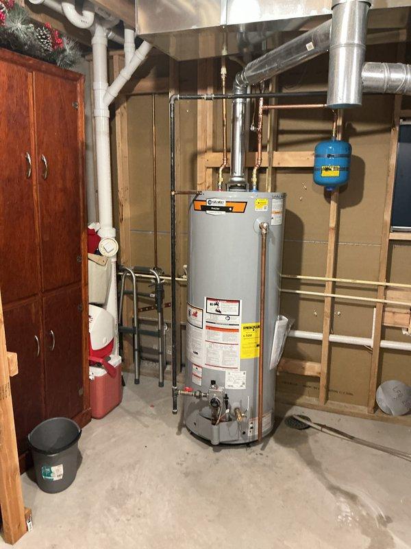 Completed professional installation of a 50-gallon tall gas water heater in an unfinished basement utility area. The State ProLine Commercial Grade unit was installed with comprehensive plumbing connections, including copper supply lines, brass fittings, multiple shut-off valves with color-coded handles for system control, and proper venting with gray flue piping. A blue thermal expansion tank was mounted to the system and secured with copper piping to protect against pressure buildup. All connections were properly fitted and sealed to manufacturer specifications, with the unit positioned on a concrete floor for optimal drainage and accessibility. The installation was completed as a cash-on-delivery setup without permit requirements.