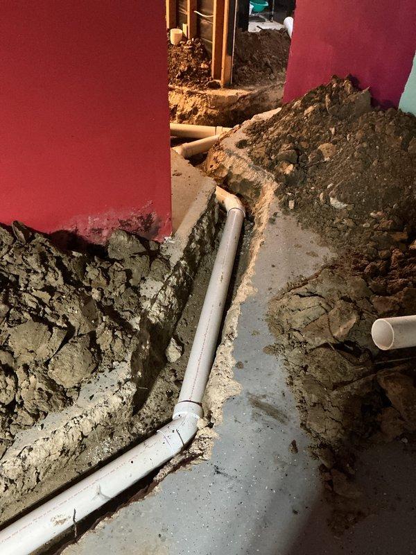Completed excavation and drainage installation with PVC DWV piping system in interior space. Trenched floor area along foundation wall, installed horizontal white PVC drain lines with proper fittings and connections, and integrated vertical gray stack pipe with coupling assembly. Applied purple primer to all PVC DWV joints per code requirements. Installation prepared for ground rough and rough-in inspection under permit 26-1648.