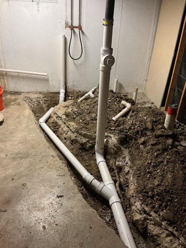 Completed excavation and drainage installation with PVC DWV piping system in interior space. Trenched floor area along foundation wall, installed horizontal white PVC drain lines with proper fittings and connections, and integrated vertical gray stack pipe with coupling assembly. Applied purple primer to all PVC DWV joints per code requirements. Installation prepared for ground rough and rough-in inspection under permit 26-1648.