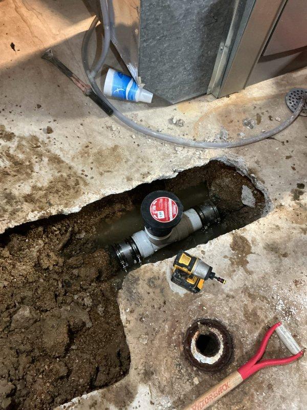 Excavated concrete floor in utility area and installed a 4-inch main drain cleanout assembly with black valve fitting and white PVC connections to provide access point for future sewer line maintenance. Backfilled excavation and ensured proper valve operation and accessibility for service purposes.
