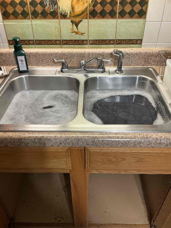 Responded to kitchen sink clog caused by food debris, specifically raspberries that had entered the drain system. Upon arrival, accessed property through back entrance as front doorbell was inoperative. Inspected double-basin stainless steel sink with standing water in both basins. Opened cabinet access panel and examined plumbing configuration, which included a Badger 500 1/2 HP garbage disposal unit connected to standard PVC P-trap drainage system. Cleared blockage from disposal and drain line, restoring proper water flow and drainage function to both sink basins.