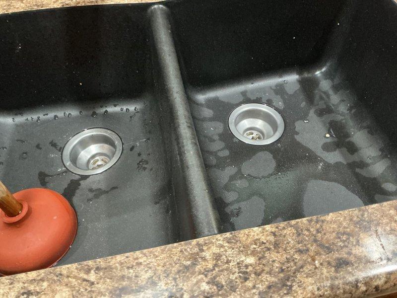 Responded to service call for clogged dual basin kitchen sink at single-family residence. Upon arrival, performed diagnostic assessment of black composite double-bowl sink with standing water and drainage issues in both basins. Inspected under-sink plumbing configuration, including white PVC P-trap assembly, drain connections, and water supply lines with braided flexible hoses and angle stop valves. Identified blockage in drain system preventing proper water flow. Utilized appropriate drain clearing tools and techniques to remove obstruction from drainage pipes. Tested both basins for proper water flow and drainage function following clearance procedure. Verified P-trap connections remained secure and leak-free. Confirmed successful restoration of full drainage capability in both sink basins. Documented conditions with photographs for service records.